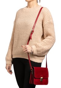 Borsa a tracolla rossa in pelle con hardware dorato, dotata di chiusura a patta e cuciture a contrasto, tenuta da una persona in un maglione beige texturizzato.