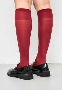Knielange rode kousen van glad materiaal met een geribbelde bovenkant, gecombineerd met glanzende zwarte schoenen met een gespdetail.