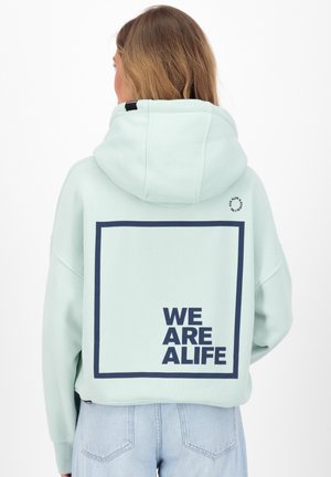 Kobieta z jasnobrązowymi włosami, ubrana w jasnoniebieską bluzę z kapturem z granatowym kwadratem i napisem „WE ARE ALIFE” na plecach, połączona z jasnoniebieskimi dżinsami.