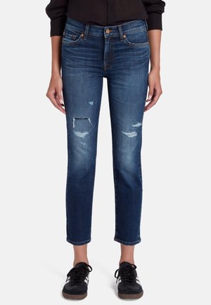 Jeans de mezclilla azules con un corte slim, que presentan detalles desgastados y dos agujeros, cinco bolsillos y una cintura de altura media, combinados con zapatillas negras.