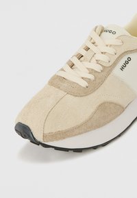 Beige suede sneaker with mesh accents, cream laces, and rubber outsole. Features "HUGO" branding on the side and a rounded toe design.