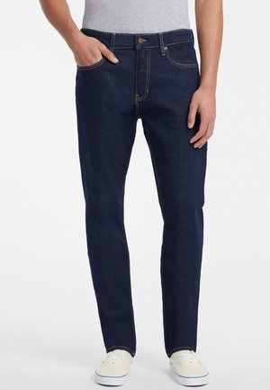 Men's dark blue denim jeans, straight fit, with yellow stitching and classic five-pocket design. Worn with a gray T-shirt and white sneakers.