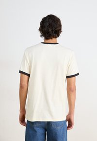 Tricou din bumbac de culoare crem cu guler și margini ale mânecilor negre. Prezintă detalii de coasere minime pe spate. Croială lejeră.