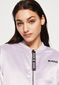 Giacca bomber di raso lilla con zip nera e branding "KANI". Presenta un collo rotondo e una texture liscia.