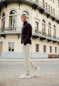 Pullover nero a coste con maniche lunghe, pantaloni a sigaretta color crema e scarpe da ginnastica bianche. In piedi su una strada lastricata vicino a un edificio vintage.