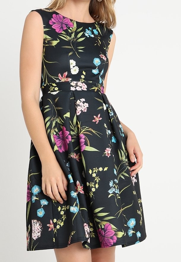 Femme portant une robe noire sans manches avec un motif floral coloré en rose, bleu, blanc et vert, debout devant un fond uni.