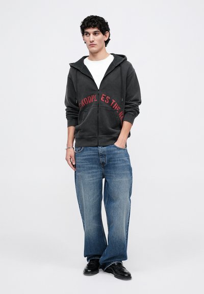 Hanorac negru cu fermoar și text roșu, purtat peste un tricou alb. Blugi largi albaștri și pantofi negri, design minimalist pe un fundal simplu.