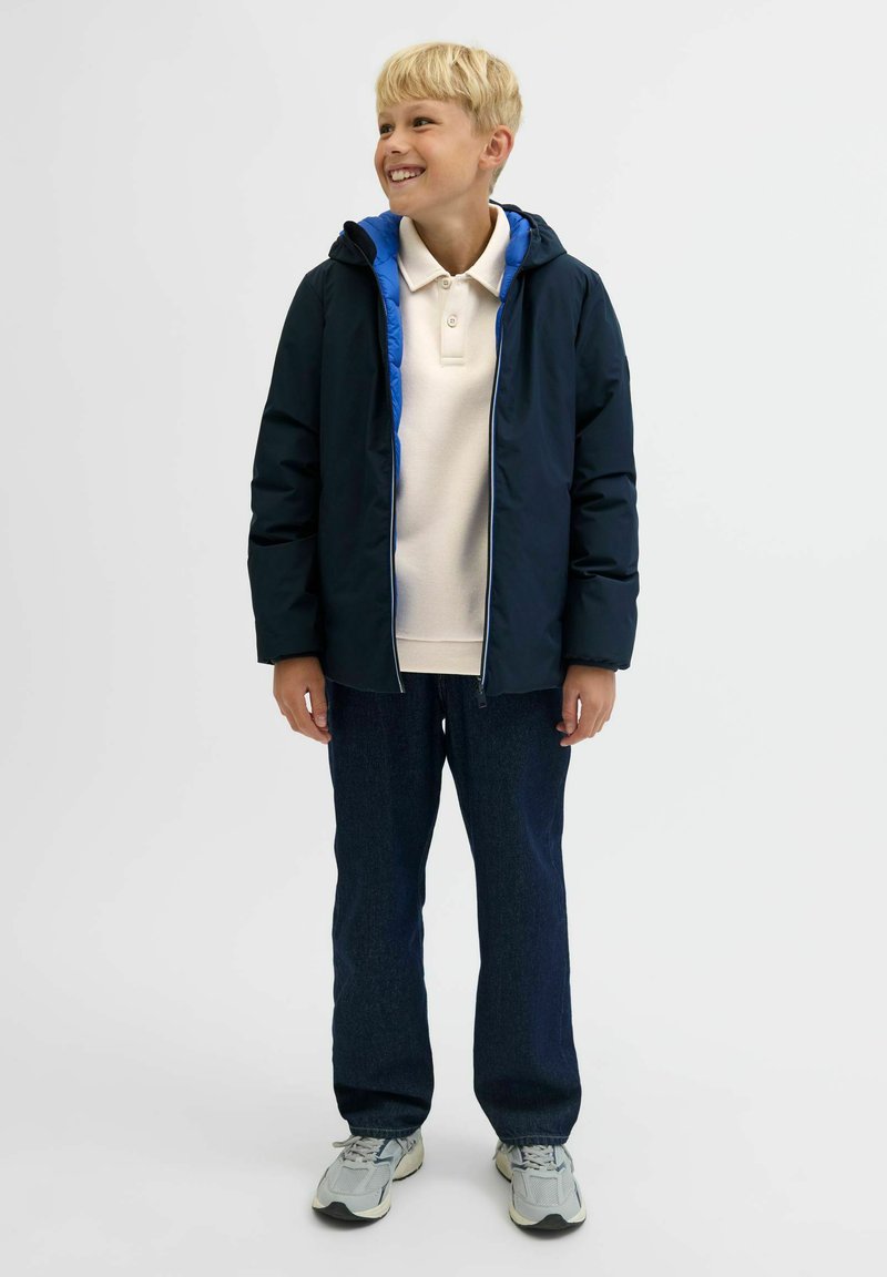 Marinblå huva jacka med blå foder, dragkedja fram; krämfärgad polo skjorta under, mörka jeans och grå sneakers. Avslappnad passform, lätta material.