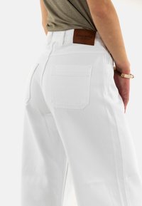 Pantalones de mezclilla blanca de pierna ancha con dos bolsillos traseros, un acabado texturizado y una etiqueta de marca de cuero marrón en la cintura.