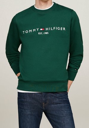 Muž nosící tmavě zelenou mikinu Tommy Hilfiger s logem a nápisem "EST. 1985", v kombinaci s modrými džíny, ruce v kapsách.