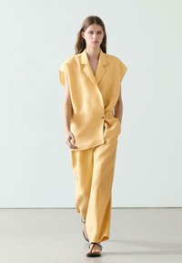 Femme portant un costume jaune ample sans manches avec un pantalon large et des sandales noires à brides, marchant devant un fond clair uni.