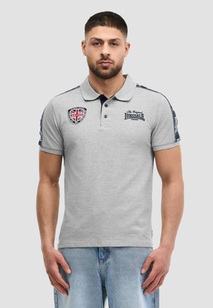 Uomo che indossa una polo grigia con toppa della bandiera britannica e logo Lonsdale London, abbinata a jeans azzurri, in piedi contro uno sfondo grigio.