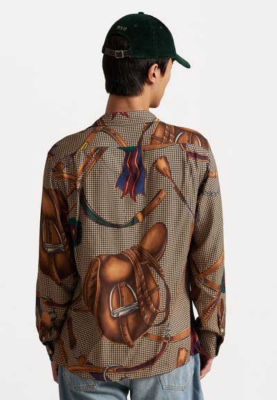 Camisa de manga larga con un estampado de silla de montar de cuero marrón sobre un fondo a cuadros. Usada con una gorra de pana verde oscura.