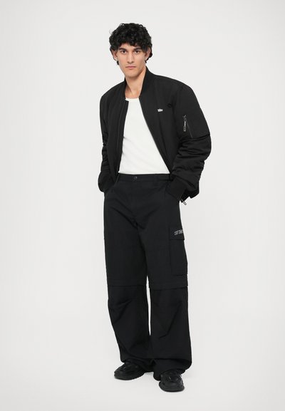 Veste bomber noire avec une poche zippée, associée à un pantalon cargo noir ample avec des poches latérales et un patch logo. T-shirt blanc en dessous.