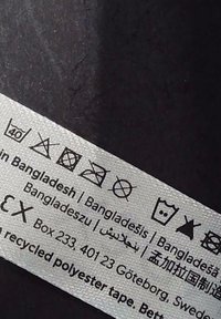 Label made of textured, woven fabric; includes care symbols and text indicating materials, origin (Bangladesh), and brand information.