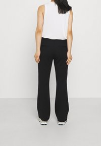 Mujer vistiendo pantalones de campana negros, una blusa sin mangas blanca y zapatillas blancas, de pie con la espalda hacia la cámara contra un fondo liso.