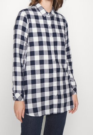 Kobieta w granatowo-białej koszuli w kratę z długim rękawem i ciemnych spodniach na jednolitym białym tle.