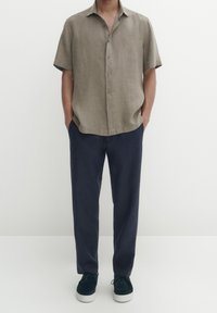 Beige linnen overhemd met korte mouwen, geknoopt, gecombineerd met donkerblauwe broek en donkergroene schoenen met witte zolen, staand tegen een neutrale achtergrond.