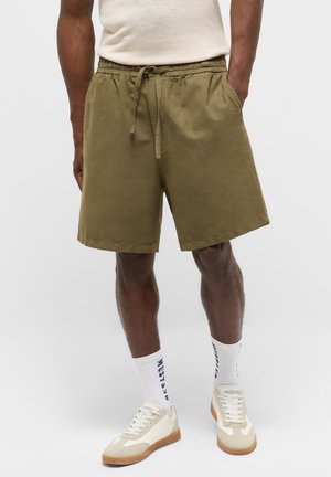 Man wearing olive green drawstring shorts, white crew socks with navy text, and beige white sneakers, standing with hands in pockets.