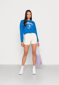 Suéter azul con texto blanco, pantalones cortos crema con dobladillo deshilachado, zapatillas blancas y una bolsa de malla lavanda. Tejido suave, ajuste casual.