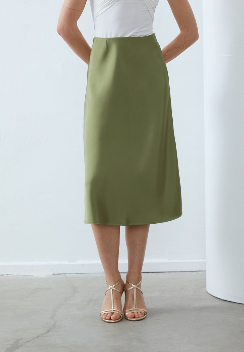 Femme portant une jupe midi taille haute olive et un haut blanc, debout avec des sandales dorées à lanières sur un sol en béton.