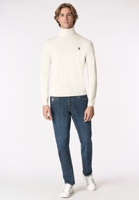Pullover bianco a collo alto con un design aderente, abbinato a jeans blu scuro. L'outfit presenta dettagli minimi e sneakers bianche.