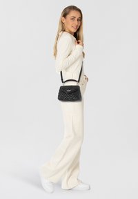 Borsa a tracolla nera trapuntata con design strutturato, hardware argento e tracolla regolabile, indossata da un modello in abbigliamento bianco. Sfondo semplice.
