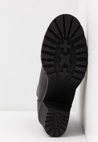 Semelle d'une botte noire avec des crampons profonds en forme de plus et de croix, positionnée contre un mur blanc d'angle.