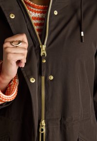 Veste brune à fermeture éclair avec tirette et boutons-pression dorés, en tissu texturé. Portée par-dessus un pull en maille multicolore avec des accents orange.