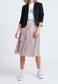 Czarna dopasowana marynarka na białym t-shircie z napisem; różowa plisowana spódnica midi; białe sneakersy z srebrnymi akcentami; gładki materiał i luźny krój.