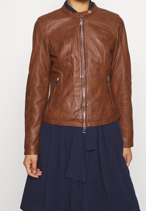 Kvinde iført en tætsiddende brun læderjakke med sølv lynlåse over en marineblå nederdel eller kjole, vist fra skuldre til hofter.