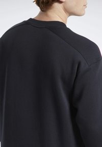 Hombre con un sudadera negra de cuello redondo simple, de espaldas, mostrando los detalles de la costura en la parte posterior y en los hombros.