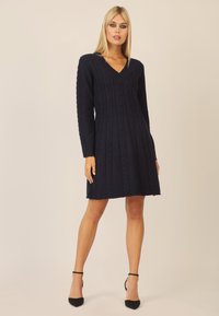 Marineblaues Strickkleid mit V-Ausschnitt, das über Kabelmuster und lange Ärmel verfügt. Der Saum endet oberhalb des Knies. Schlicht mit minimalen Akzenten.