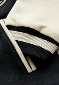Suéter negro y crema con puños acanalados y un diseño de rayas audaces, fabricado en material de algodón suave y con costuras detalladas.