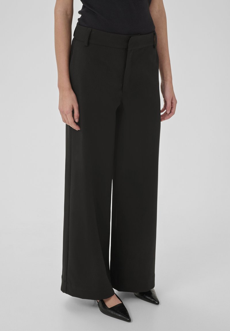 Personne portant un pantalon noir à jambes larges et des chaussures noires à bout pointu, debout devant un fond gris uni.