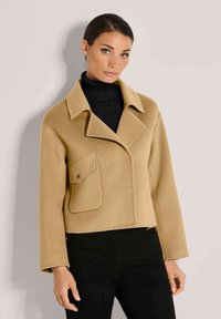 Frau in einem kurzen, camel-farbenen Mantel mit einer großen Vorderasche und breitem Kragen über einem schwarzen Rollkragenpullover und schwarzen Hosen.