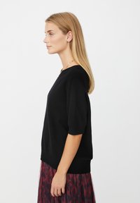 Kvinde med glat, blond hår iført en sort kortærmet sweater og en rød mønstret nederdel, stående i profil mod en hvid baggrund.