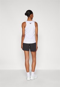 Top blanco sin mangas con un logo en la parte posterior, combinado con pantalones cortos oscuros y zapatillas blancas. El fondo es de un gris claro liso.