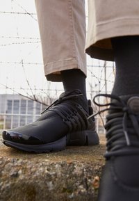 Osoba oblečená v černých teniskách a černých ponožkách, stojící na kamenném povrchu s drátěným plotem a budovou v pozadí.