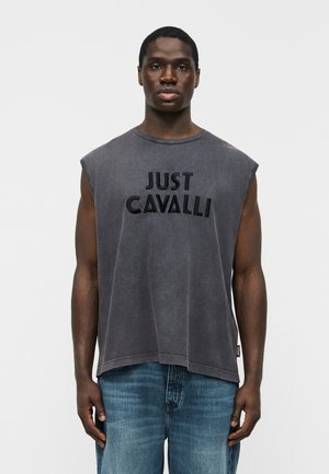 Bărbat purtând un tricou fără mâneci, negru șters, cu textul "JUST CAVALLI" și blugi albaștri, stând pe un fundal alb simplu.