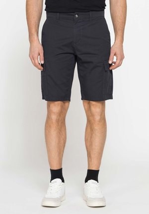 Uomo che indossa pantaloncini cargo neri, calzini neri e sneakers bianche, in piedi davanti a uno sfondo chiaro e uniforme.