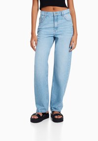 Jeans de tiro alto y pierna recta en azul claro, combinados con sandalias de plataforma negras y una blusa negra sin mangas que deja al descubierto el abdomen y los tobillos.