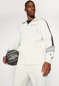 Sudadera con cremallera de color crema claro con detalles en negro y gris, bolsillos frontales y logo de Champion. El modelo sostiene un balón de baloncesto negro.