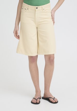 Osoba oblečená v béžových krátkych nohaviciach po kolená, zelenom rebrovanom tričku a čiernych žabkách, stojaca na jednoduchom svetlom pozadí.