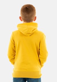 Sudadera con capucha amarilla de textura suave, con puños y dobladillo acanalados. La vista trasera muestra las costuras y el pliegue en la capucha. Llevada sobre jeans azules.