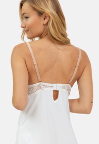 Camisole blanche avec dentelle au dos, dotée de bretelles nude réglables et d'une petite ouverture en forme de clé. Texture de tissu lisse.
