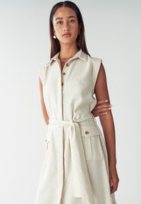 Giovane donna con lunghi capelli scuri, vestita con un abito beige senza maniche, con cintura, bottoni e taschini, in piedi davanti a uno sfondo neutro.