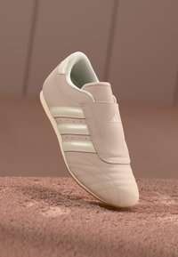 Bežs Adidas sporta apavi ar baltām svītrām un logo, novietoti uz teksturētas rozā virsmas pret fona rozā.