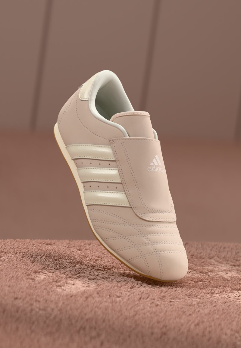 Zapatilla Adidas beige con franjas y logo blancos, colocada sobre una superficie texturizada rosa contra un fondo rosa suave.