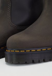 Gros plan sur des bottes en cuir brun foncé avec panneaux latéraux élastiques noirs, coutures jaunes et semelles épaisses rainurées noires.